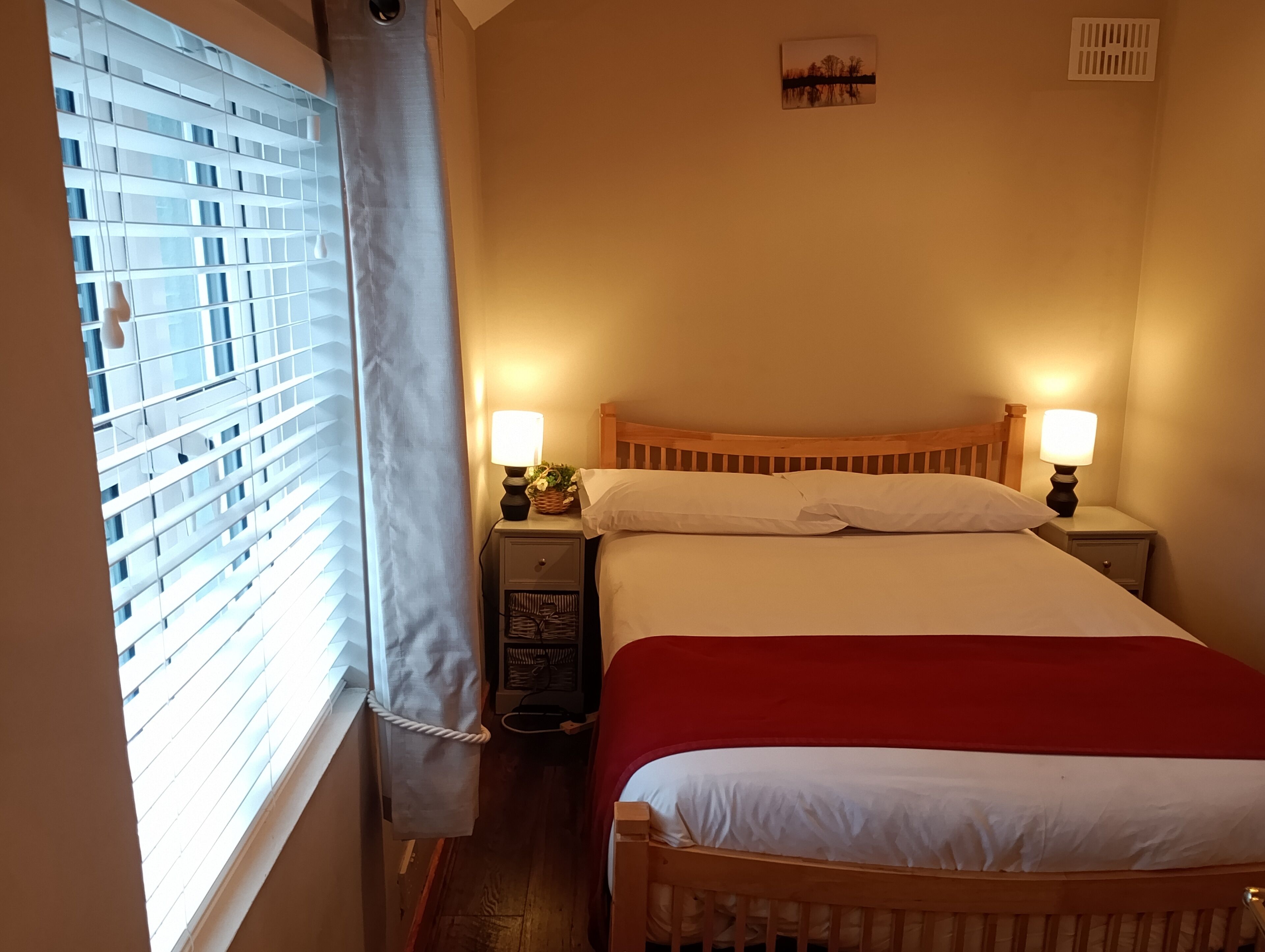 A bedroom with a wooden bed, two bedside tables with lamps, a nightstand, a window with blinds, and a small picture on the wall.