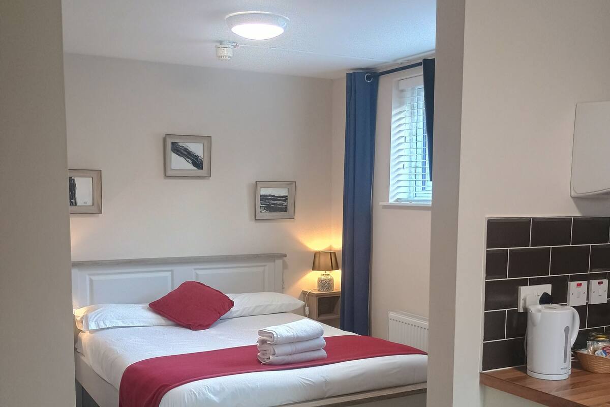 A hotel room with a bed, a nightstand, a lamp, a window with curtains, a radiator, a tiled backsplash, and a trash bin.
