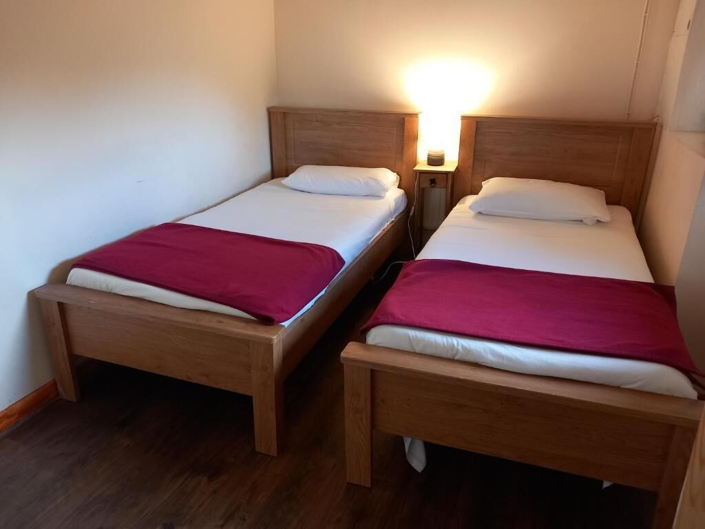 Dos camas de madera con ropa de cama blanca y roja, una mesita de noche con lámpara y suelo de madera.