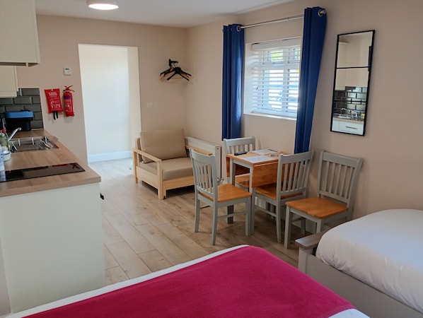 A compact room with a kitchenette, a dining table with chairs, a bed, and a window with curtains.