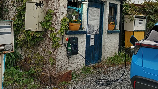 Un petit bâtiment avec une porte bleue, une horloge et une borne de recharge pour une voiture bleue.
