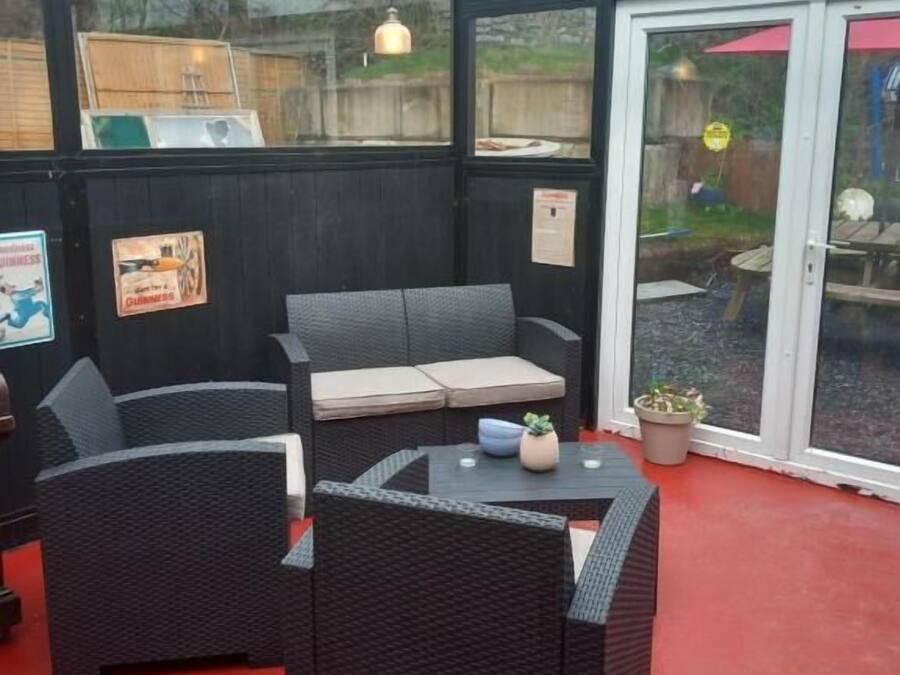 En overdækket terrasse med fletningsmøbler, herunder en sofa og to stole, et lille bord med en plante, og en glasdør til haven.