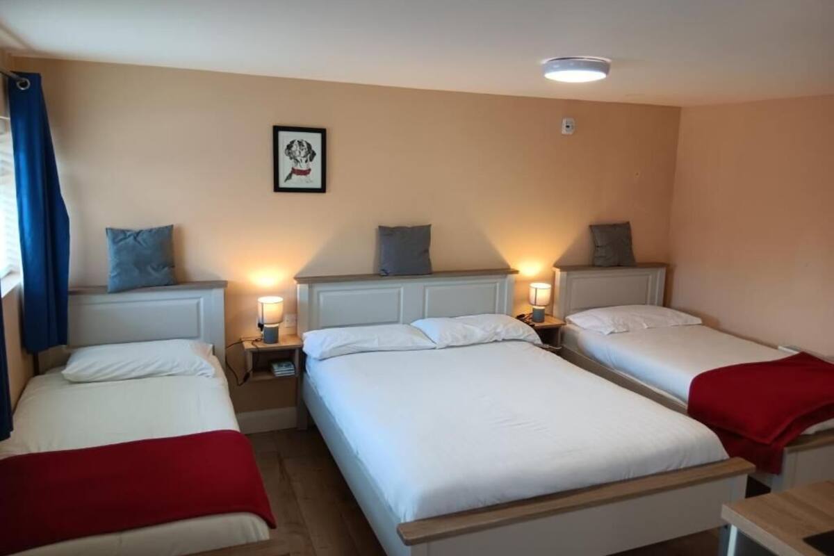 A hotel room with three single beds, each with a bedside table and lamp. The room has a neutral color scheme with beige walls and white bedding.