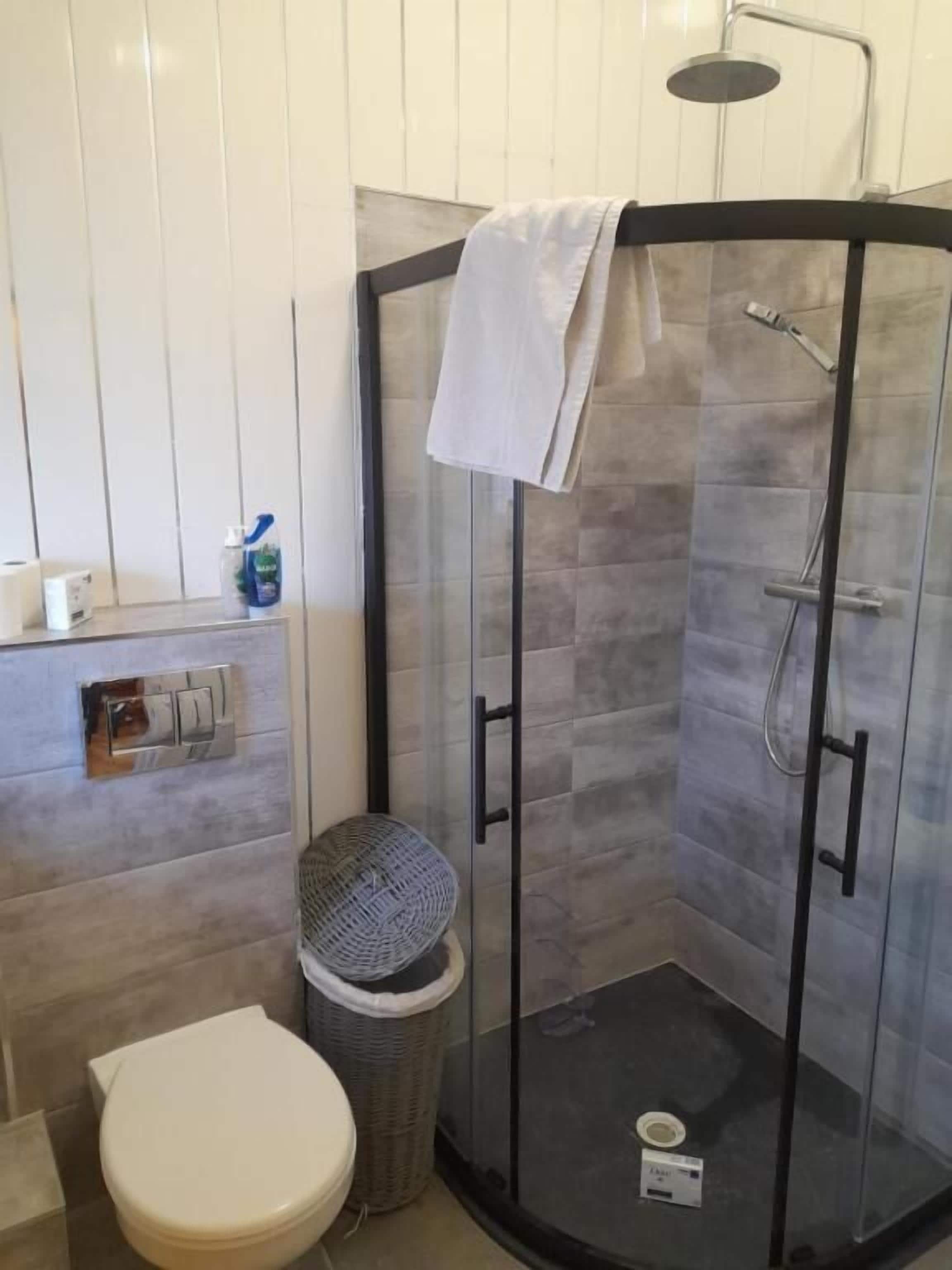 Un baño con ducha de vidrio, un inodoro, un cubo de basura y una toalla colgada en la puerta de la ducha.