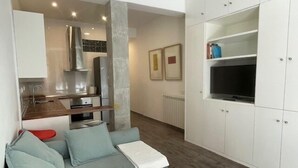 Een moderne woonkamer met een grijze bank, een witte salontafel, en een keukenhoek met witte kasten en een inbouwoven.