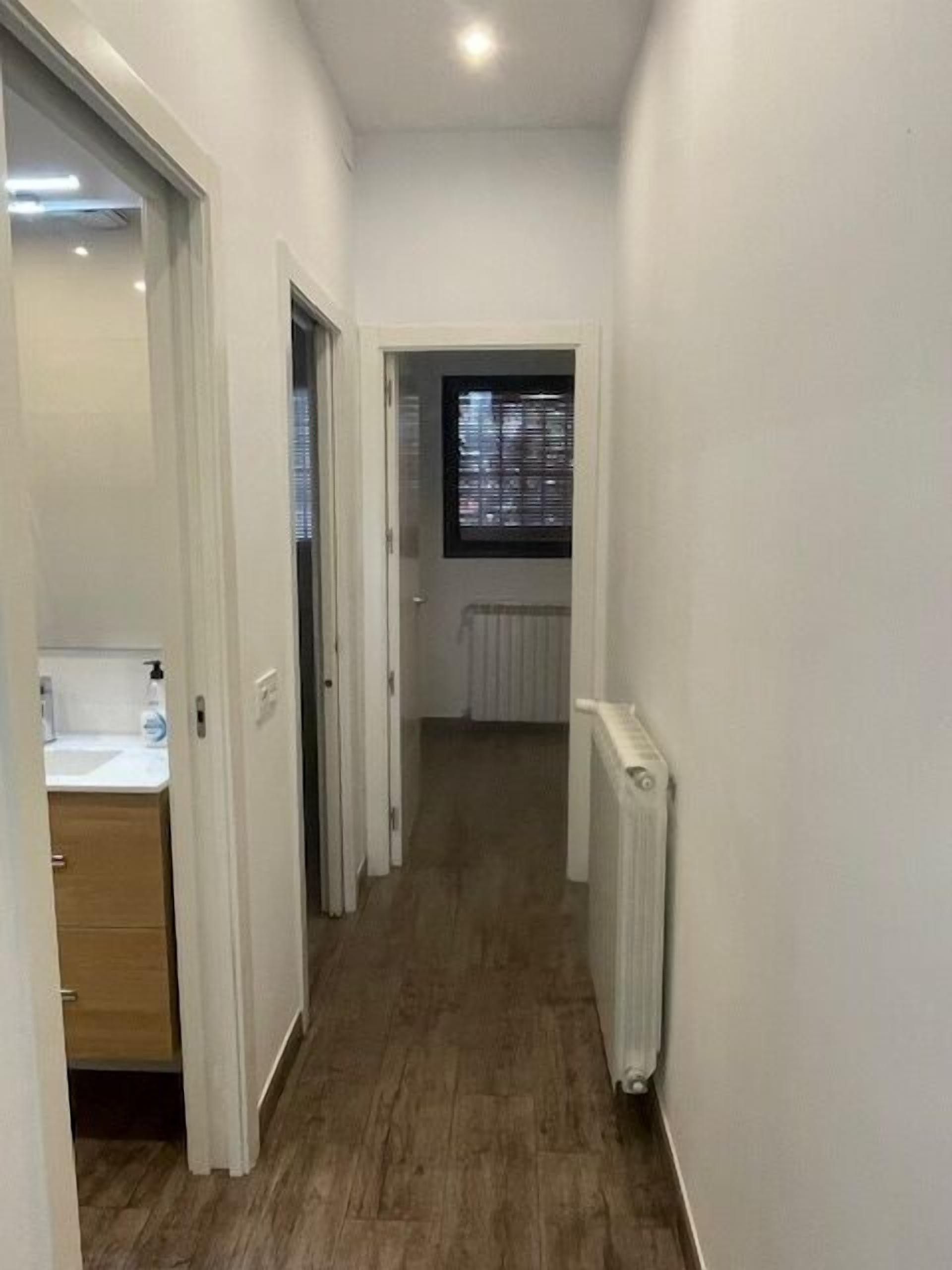 A narrow hallway with wooden flooring, white walls, and a window with blinds.