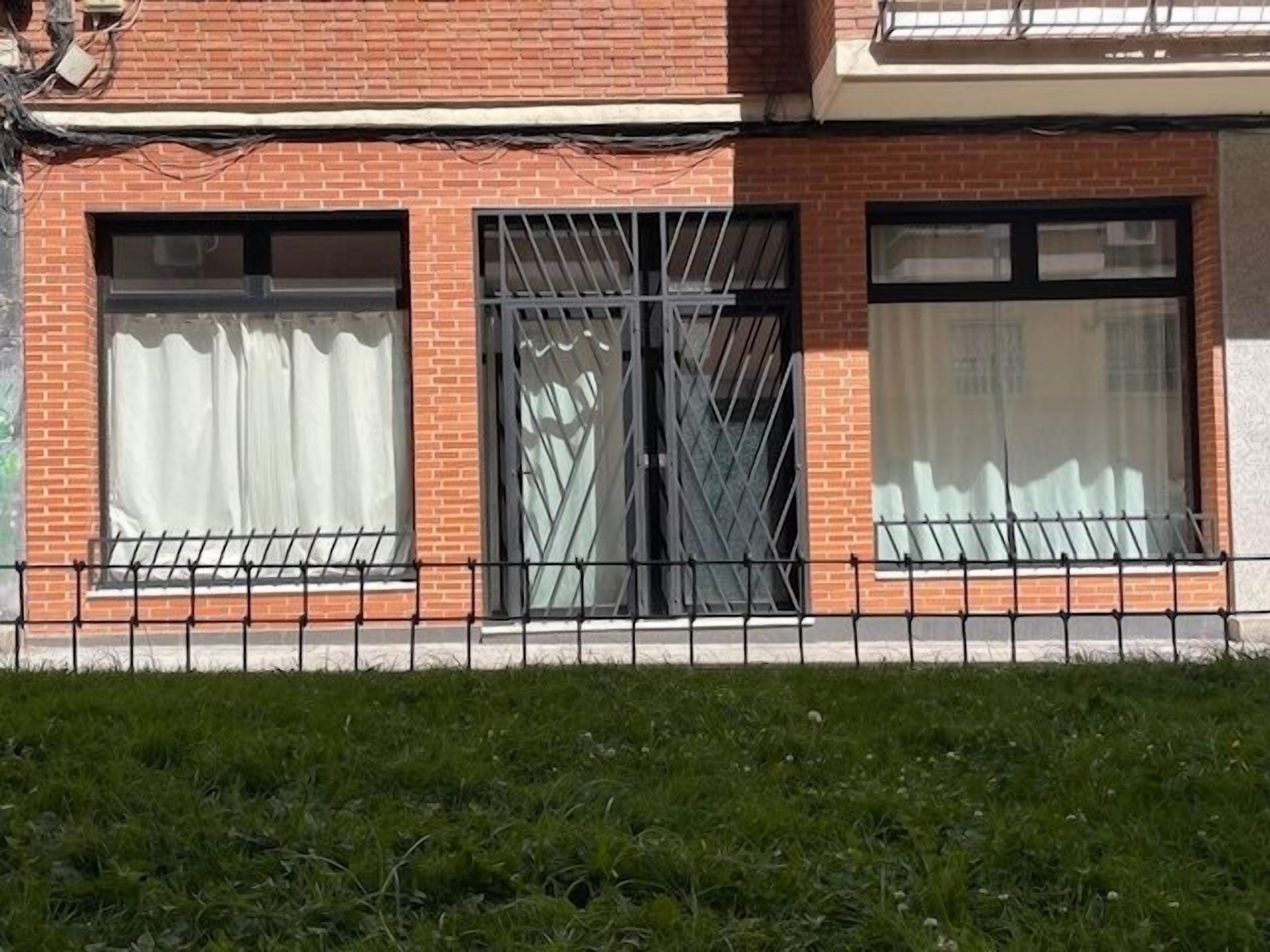 A brick building with three large windows, one with a metal grille, and a black metal fence in front.