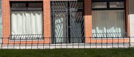 A brick building with three large windows, one with a metal grille, and a black metal fence in front.
