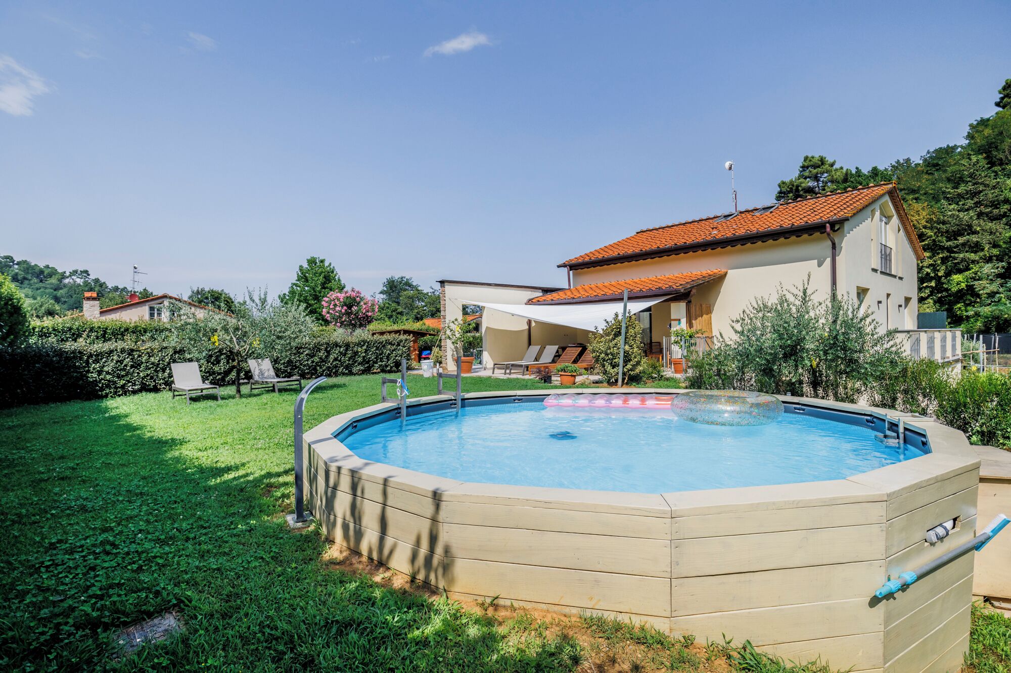 A swimming pool with a wooden deck, surrounded by a well-maintained lawn and a house with a red-tiled roof.