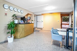 Zona de recepción con mostrador, relojes, planta en maceta y un área de estar con mesas y sillas.