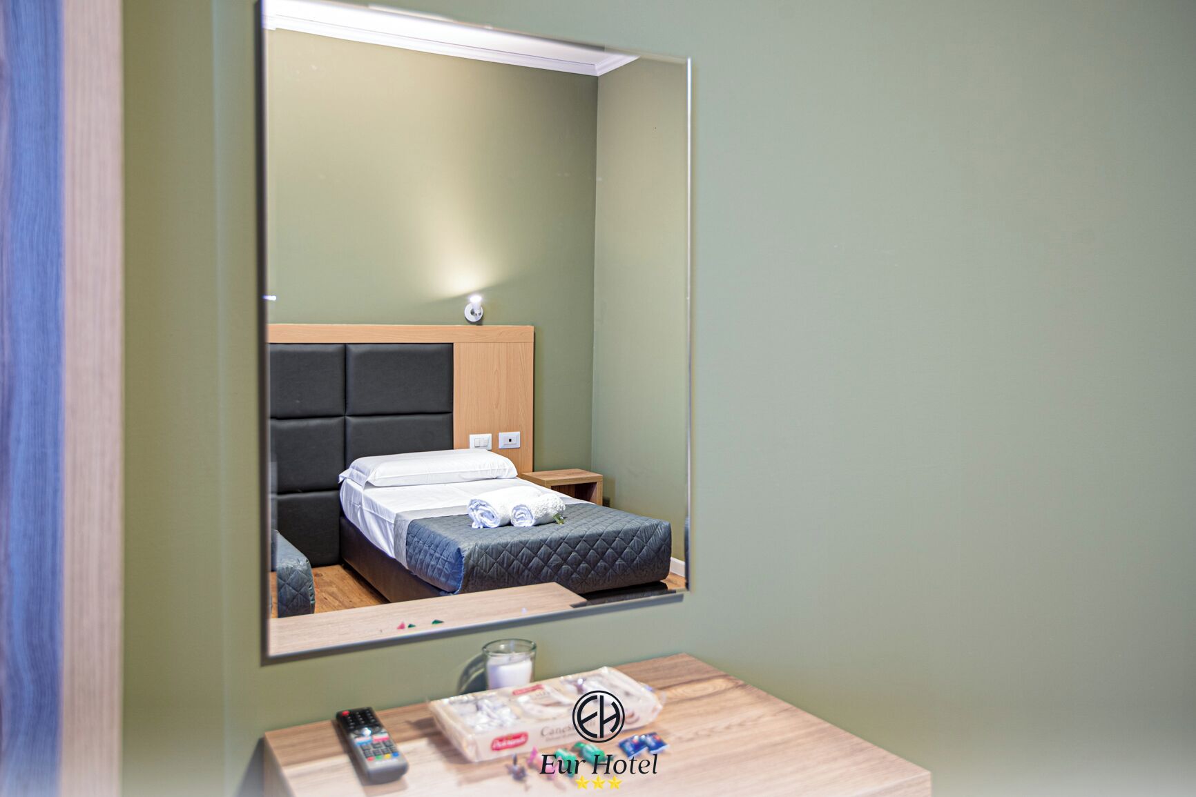 A hotel room with a bed, a bedside table with a remote control, a glass of water, and toiletries.