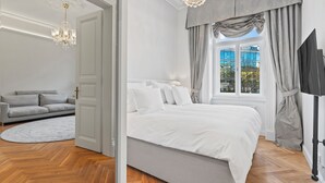 A bedroom with a bed, a grey sofa, a chandelier, and a window with curtains. - Vodičkova Luxury Suites (Prague)