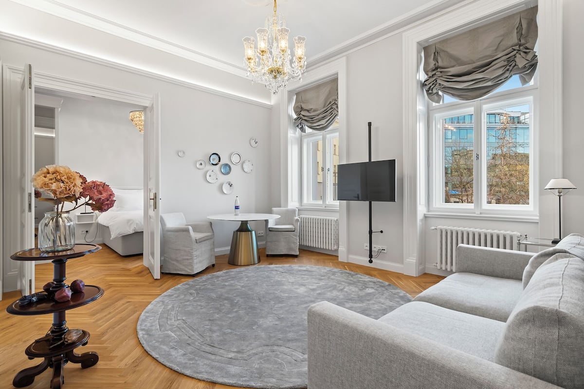 Un salon moderne avec un tapis gris, des meubles blancs, un lustre et un téléviseur fixé au mur.
