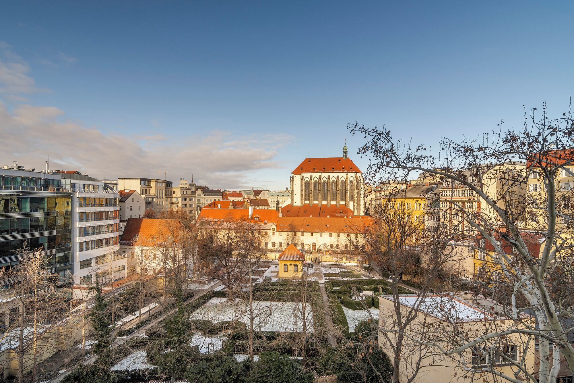 A historic building with a red roof, surrounded by modern buildings and a snow-covered garden.