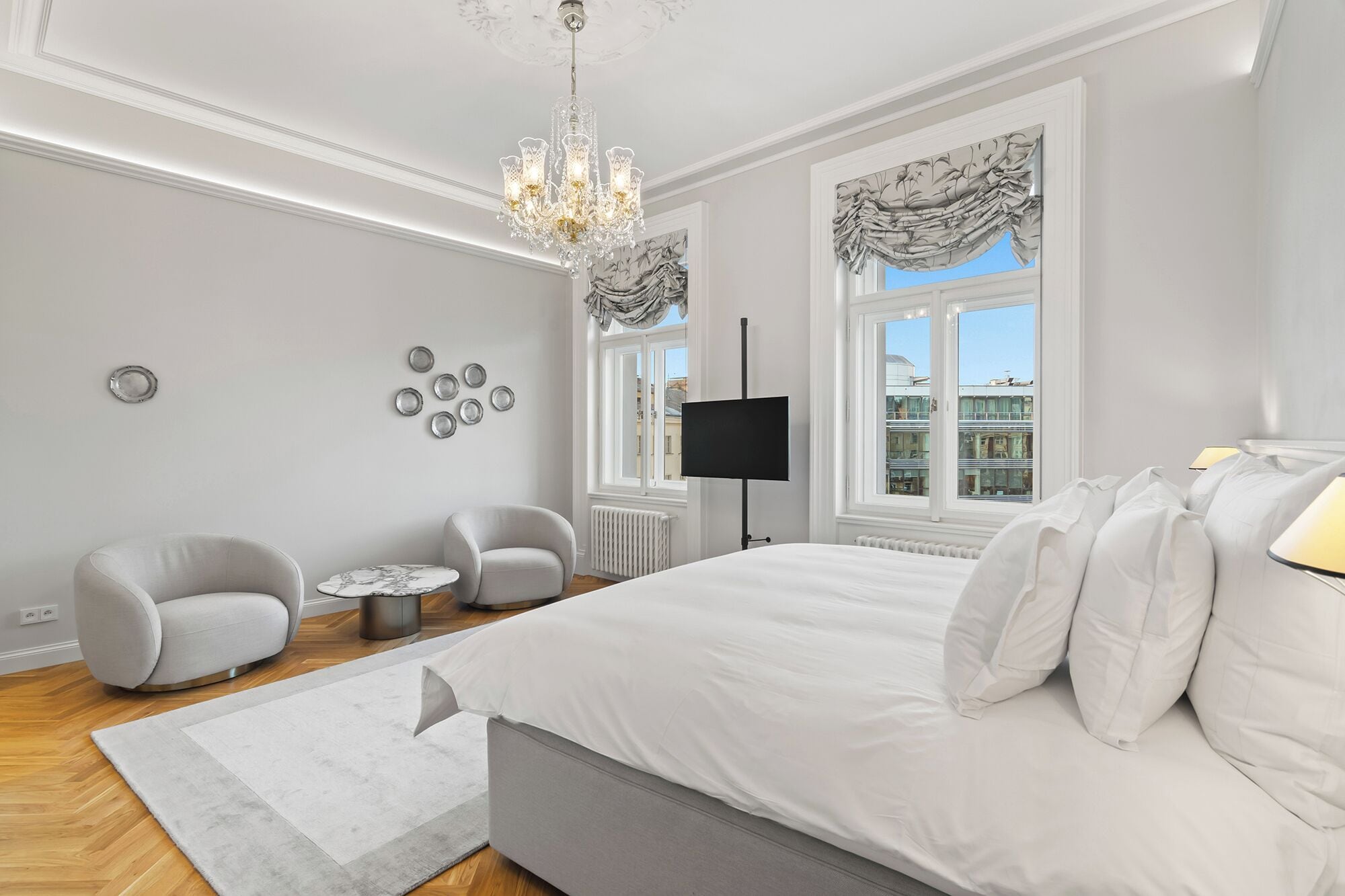 A modern bedroom with a large bed, two armchairs, a small table, a chandelier, a TV, and a window with a view of buildings.