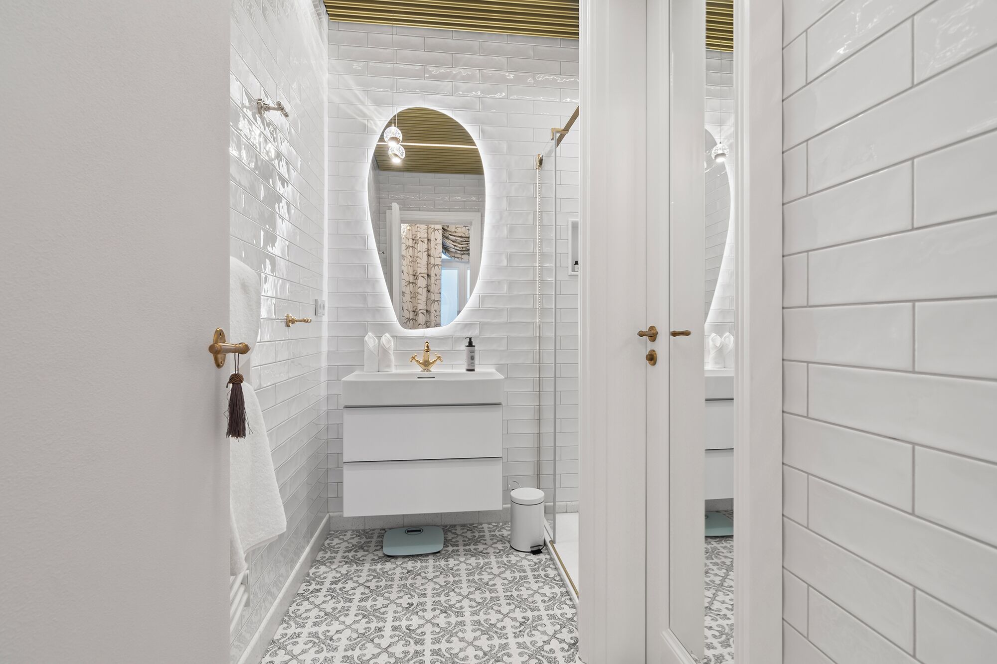 A narrow bathroom with white tiled walls, a white sink with gold fixtures, and a patterned floor.