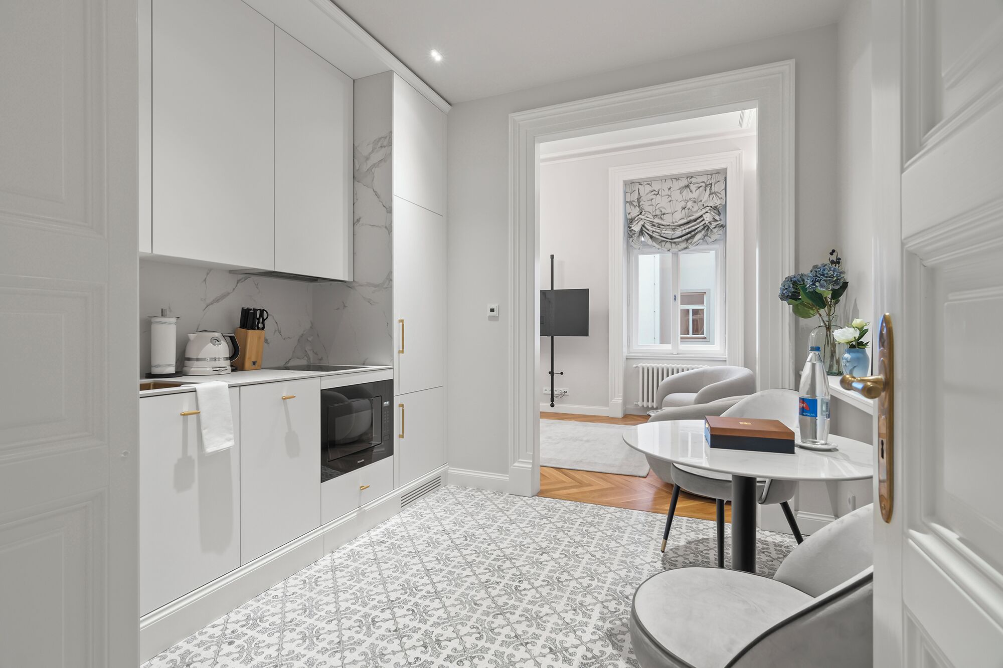 A modern kitchen with white cabinetry, a built-in oven, and a coffee machine. There is a dining area with a round table and chairs, and a view of a living room through an open door.