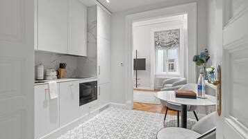 A modern kitchen with white cabinetry, a built-in oven, and a coffee machine. There is a dining area with a round table and chairs, and a view of a living room through an open door.