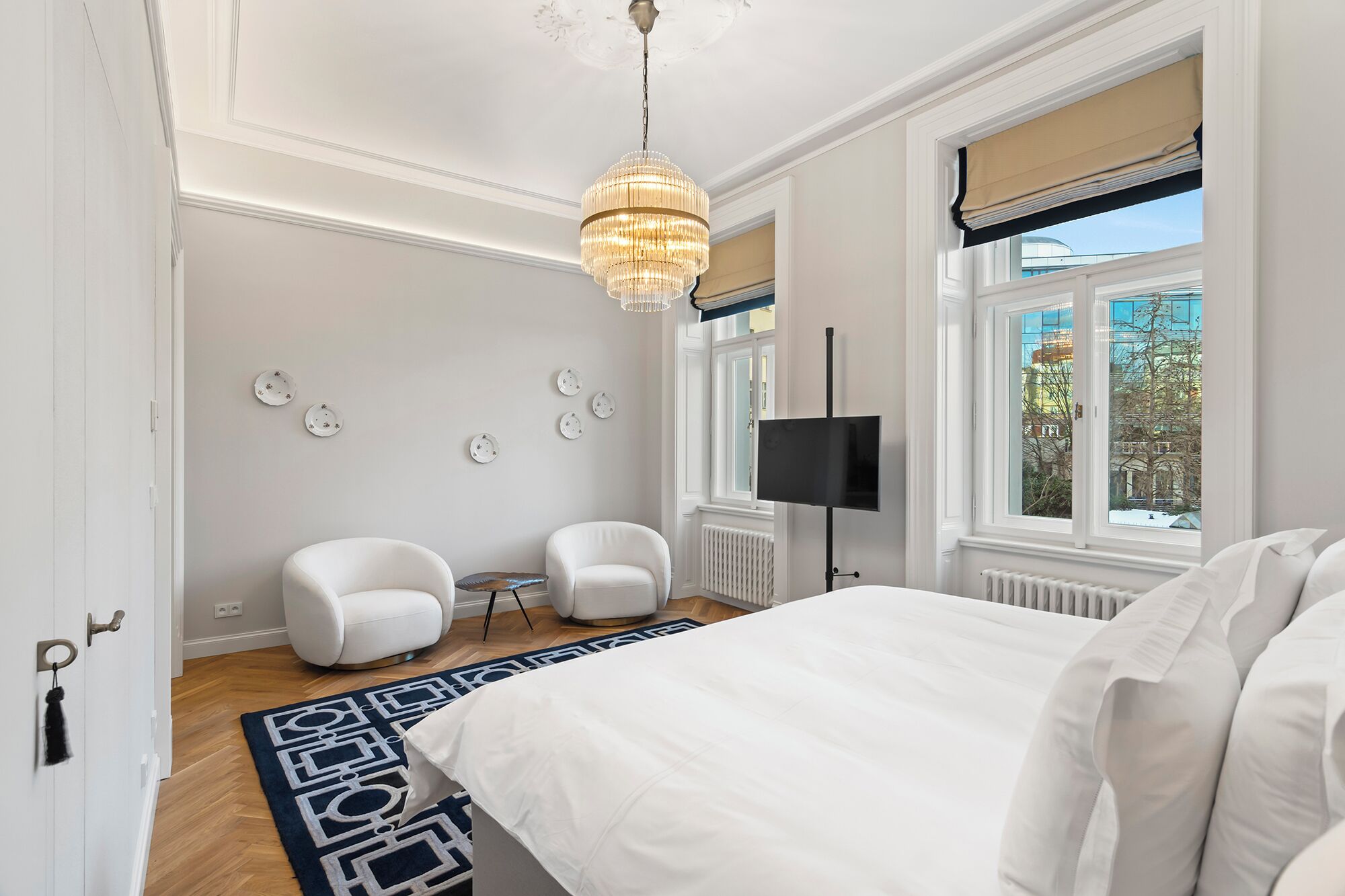 A bedroom with a large bed, two white armchairs, a chandelier, a TV, and a window with a view of buildings.