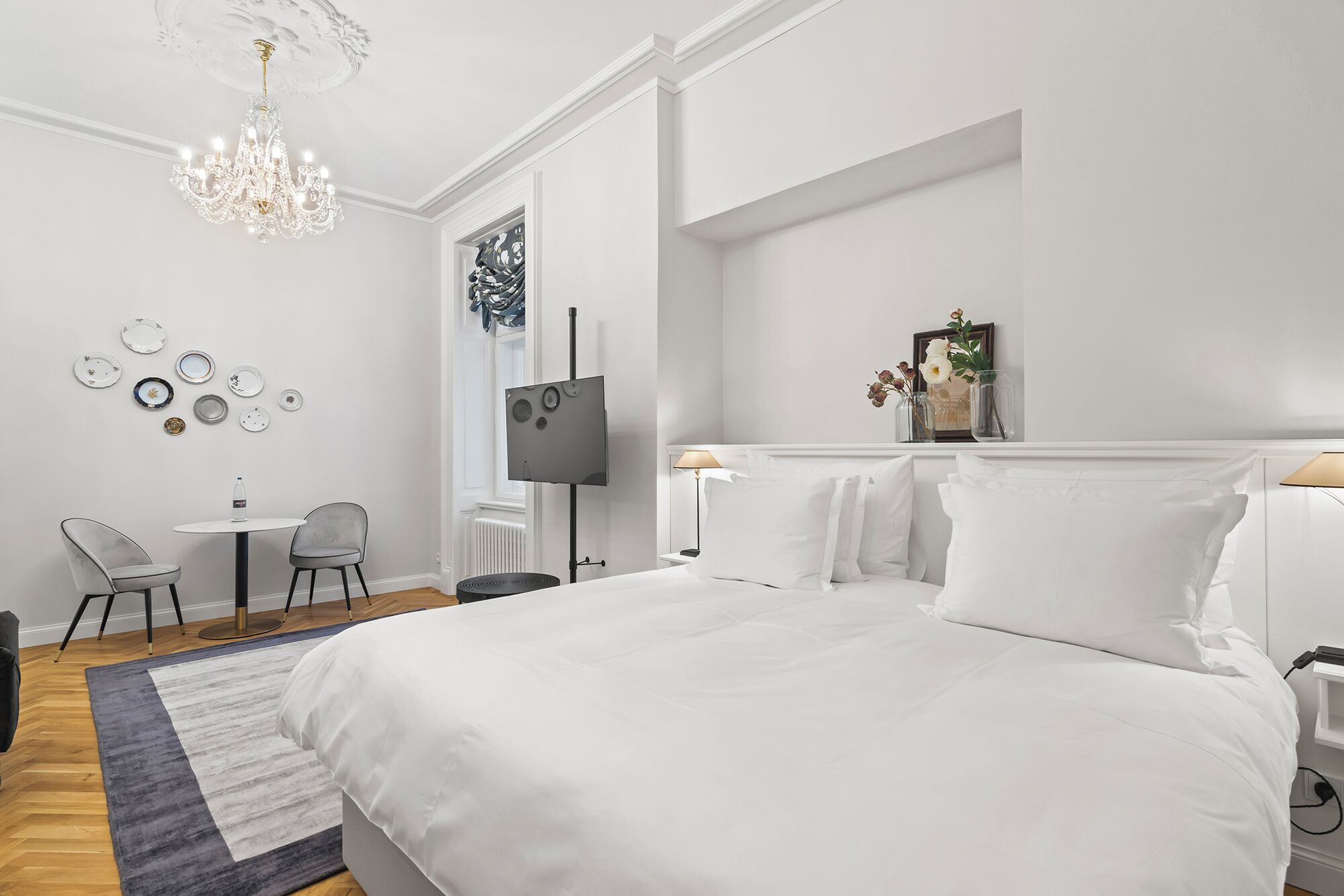 Une chambre d’hôtel moderne avec un grand lit, un lustre, un téléviseur à écran plat, une petite table avec des chaises et un téléviseur fixé au mur.