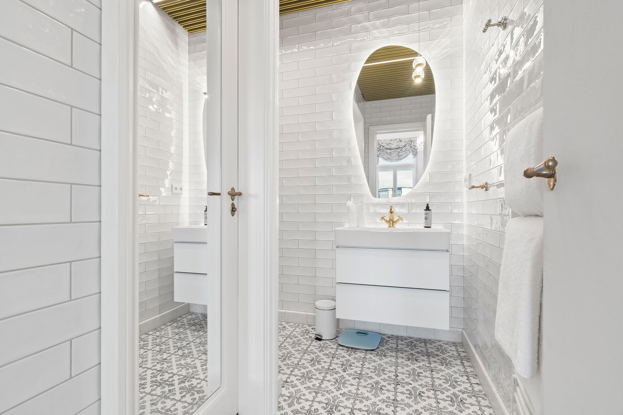 Un baño moderno con azulejos blancos tipo metro, un tocador blanco con detalles dorados y baldosas de piso con diseño.