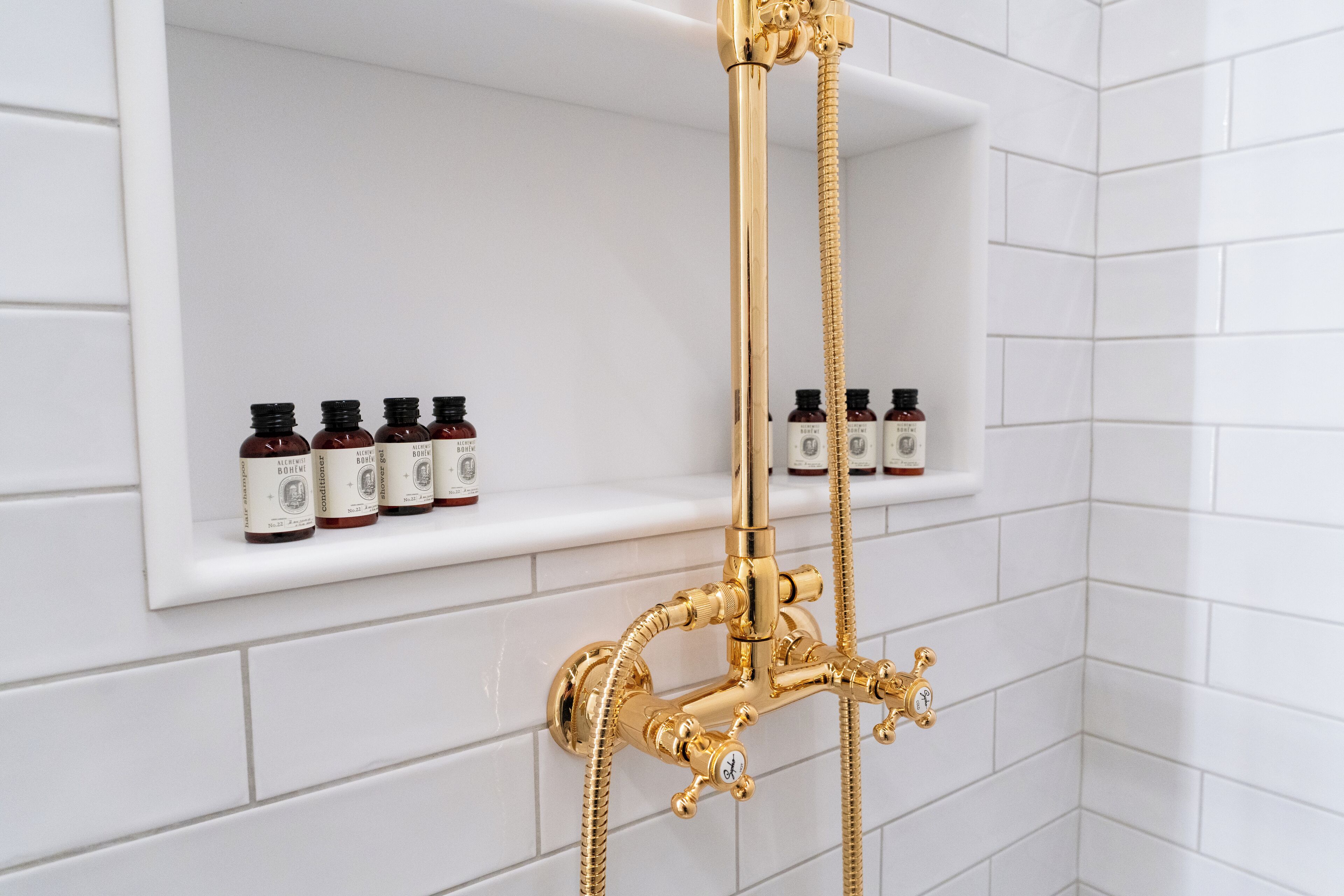 A bathroom with a gold showerhead and control valves, a white tiled wall, and a shelf with several bottles.