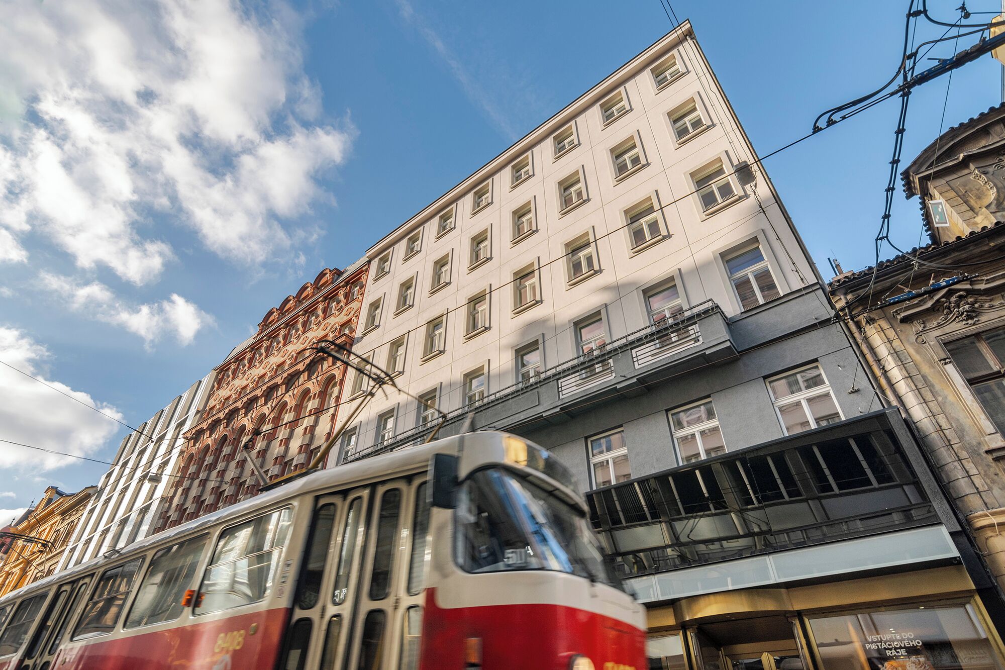 A tram moving past a building with a classical facade and a modern lower section.