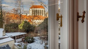 A view through a window of a building with a red roof and a tower, with snow on the ground. - Vodičkova Luxury Suites (Prague)