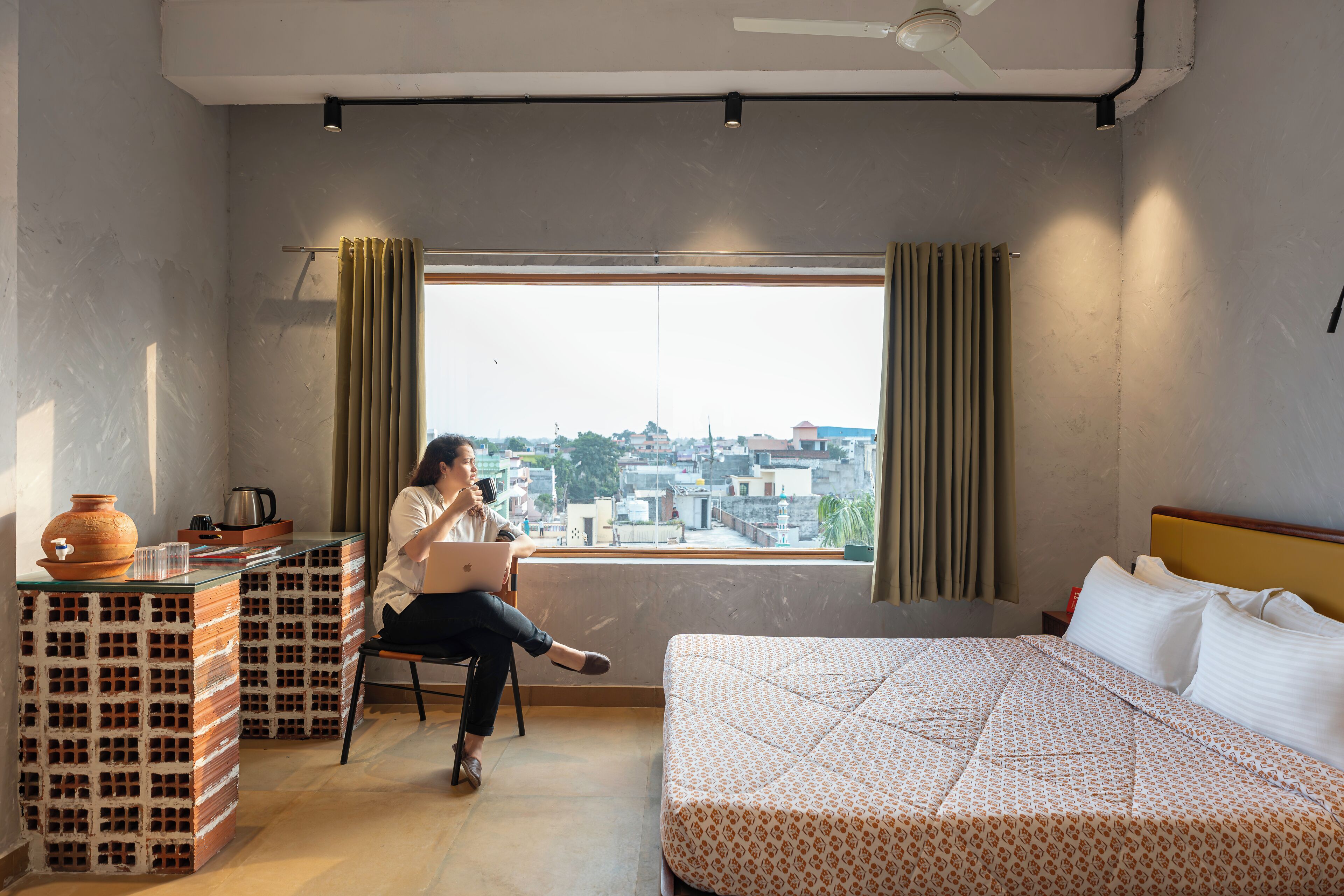 A room with a bed, a desk with a laptop, a window with curtains, and a view of the city.