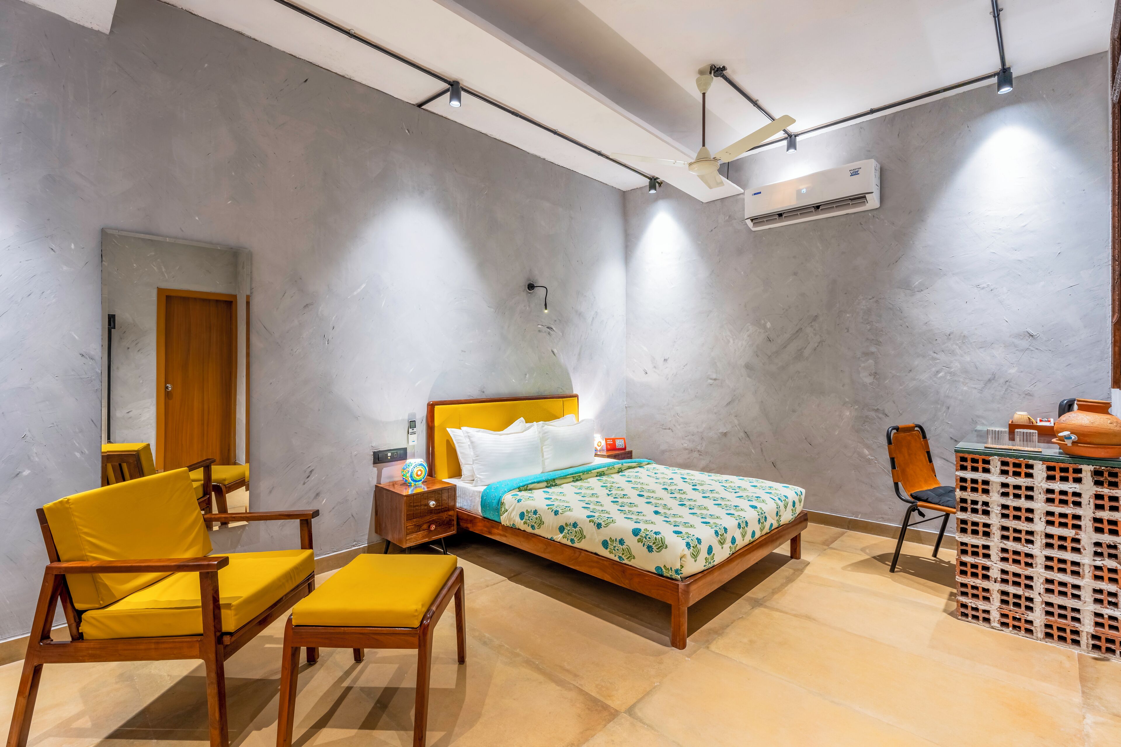 A bedroom with a bed, a wooden nightstand, a chair, a small table, and an air conditioning unit.