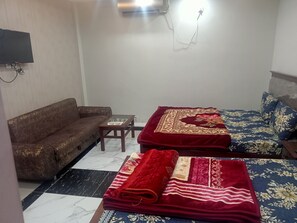 A small, furnished room with a sofa, two beds with floral bedding, a small table, and a TV mounted on the wall. - Four Season Guest House (Islamabad)