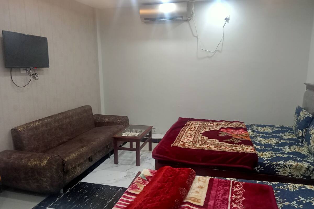 A room with a sofa, two beds with red and blue floral bedspreads, a small table, and a mounted TV.