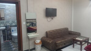 Een woonkamer met een bank, een klein tafeltje, een prullenbak, een aan de muur bevestigde televisie en een badkamer die door een open deur zichtbaar is.