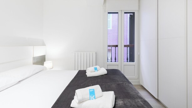 A modern hotel room with a large bed, a bedside table, a radiator, and a window with a view of a building.
