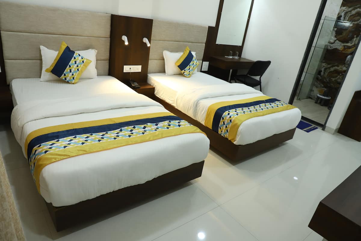 A hotel room with two beds, each with a patterned bedspread, a bedside table with a lamp, and a small desk with a chair.