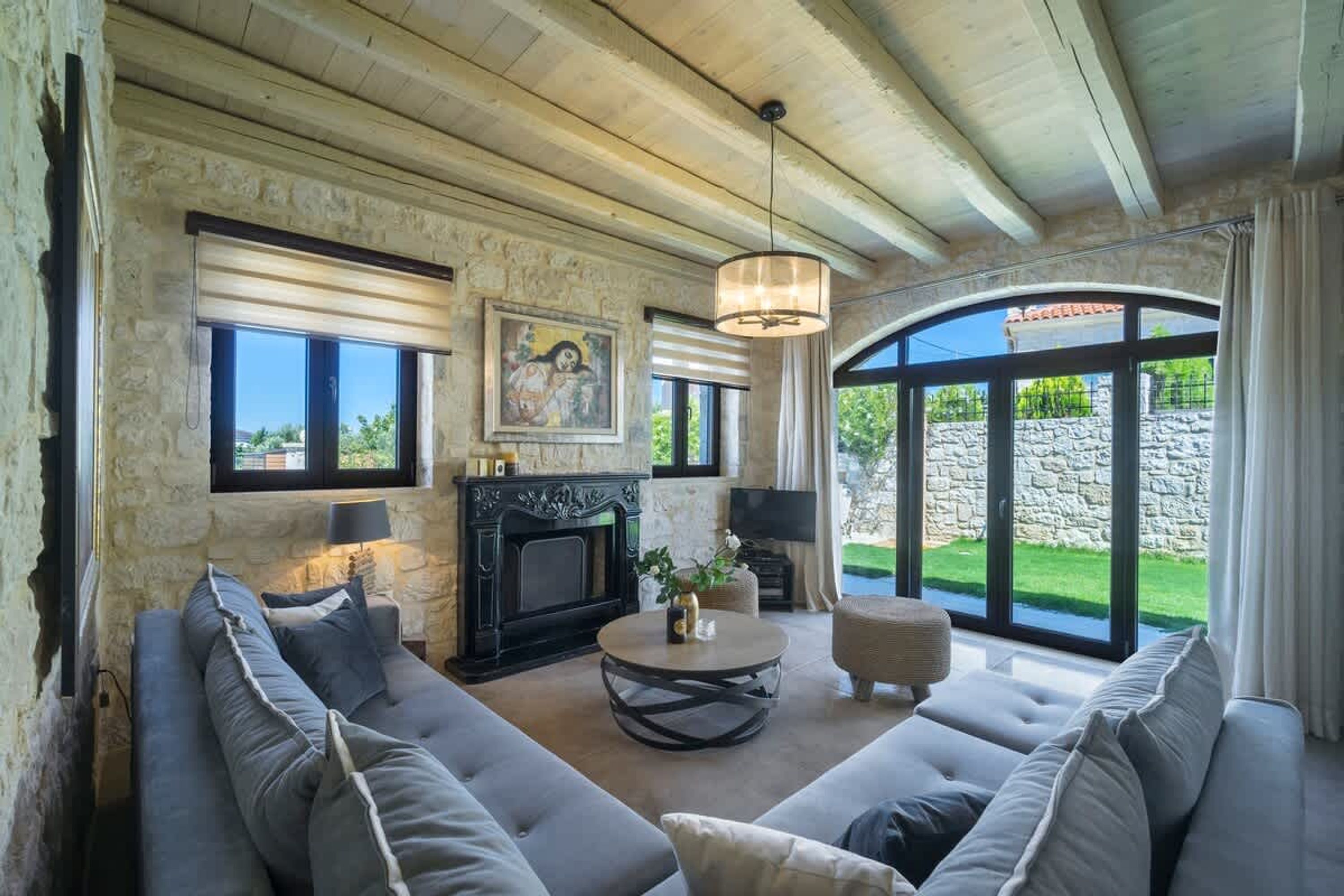A spacious living room with a stone fireplace, a large sofa set, a round coffee table, and a large window offering a view of the outdoors.