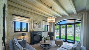 A spacious living room with a stone fireplace, a large sofa set, a round coffee table, and a large window offering a view of the outdoors. - Heraklion Twins House - Private Pool Retreat (Hersonissos)