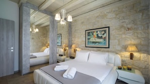 A modern hotel room with a stone wall, a large bed with white linens, a mirror, and a chandelier. - Heraklion Twins House - Private Pool Retreat (Hersonissos)
