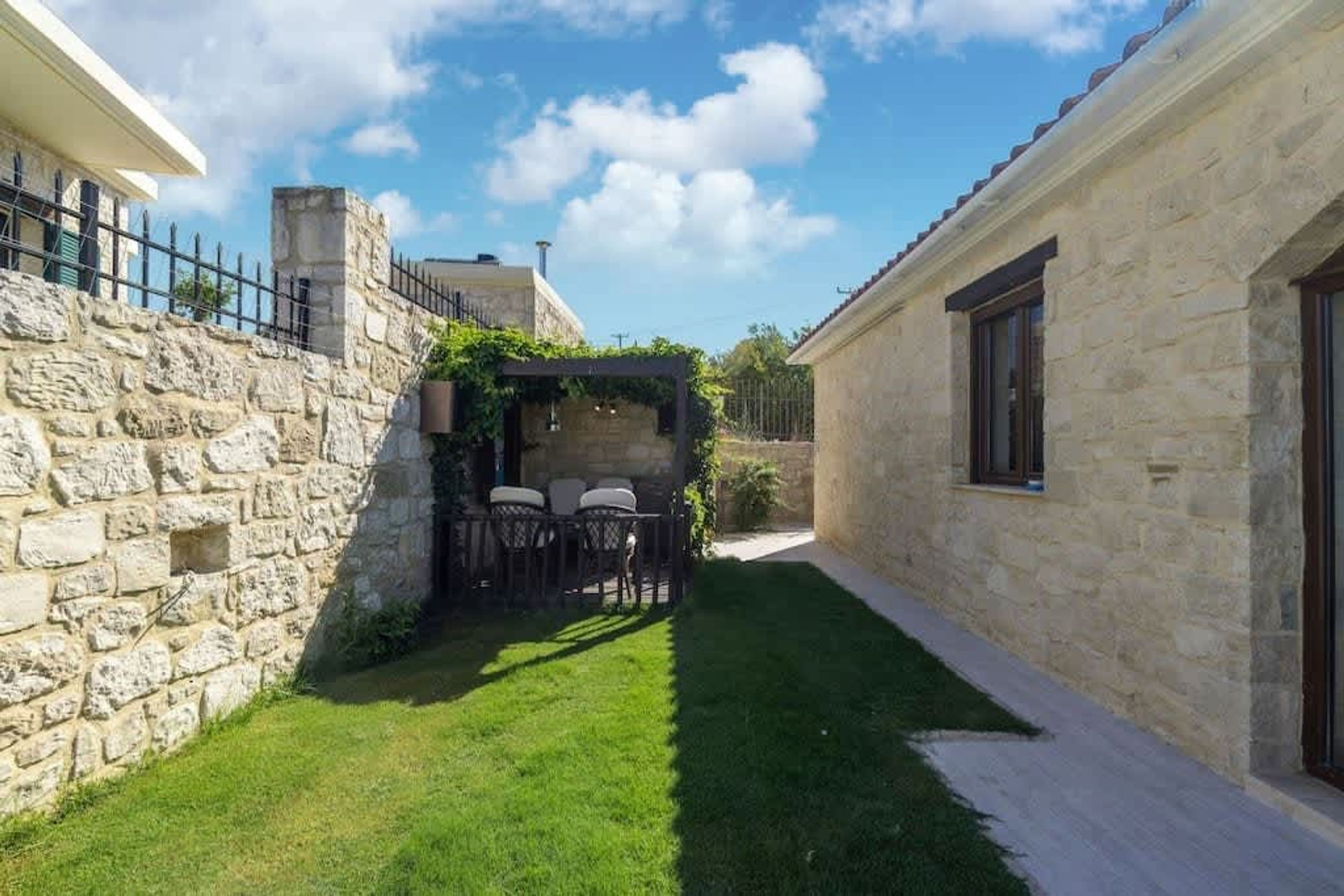 A narrow courtyard with a stone wall on one side and a building on the other. There is a small seating area with a table and chairs.