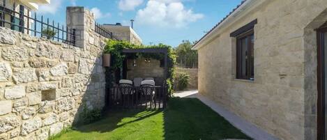 A narrow courtyard with a stone wall on one side and a building on the other. There is a small seating area with a table and chairs.
