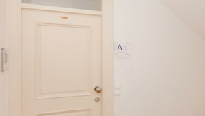 Egy fehér ajtó, felette egy kis ablak, egy fa lépcső, melynek alján barna szőnyeg van, és a falra szerelt tábla, melyen az „AL” betűk láthatók.