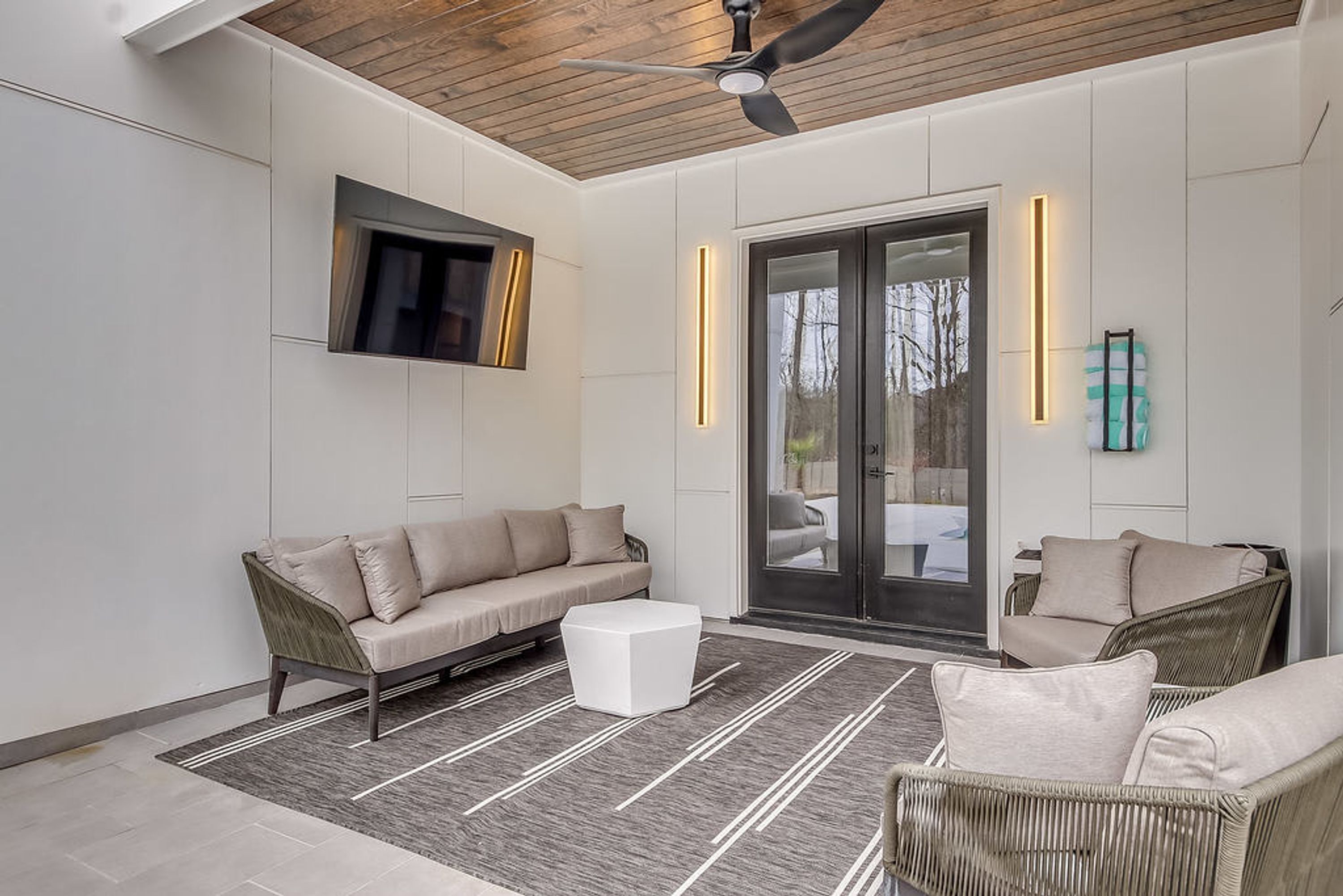 A modern living room with a sofa, a coffee table, and wicker chairs. There is a ceiling fan, a flat-screen TV mounted on the wall, and a sliding glass door leading outside.