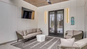A modern living room with a sofa, a coffee table, and wicker chairs. There is a ceiling fan, a flat-screen TV mounted on the wall, and a sliding glass door leading outside.