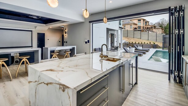 Una cocina moderna con una isla de mármol, iluminación colgante y vista a una piscina.