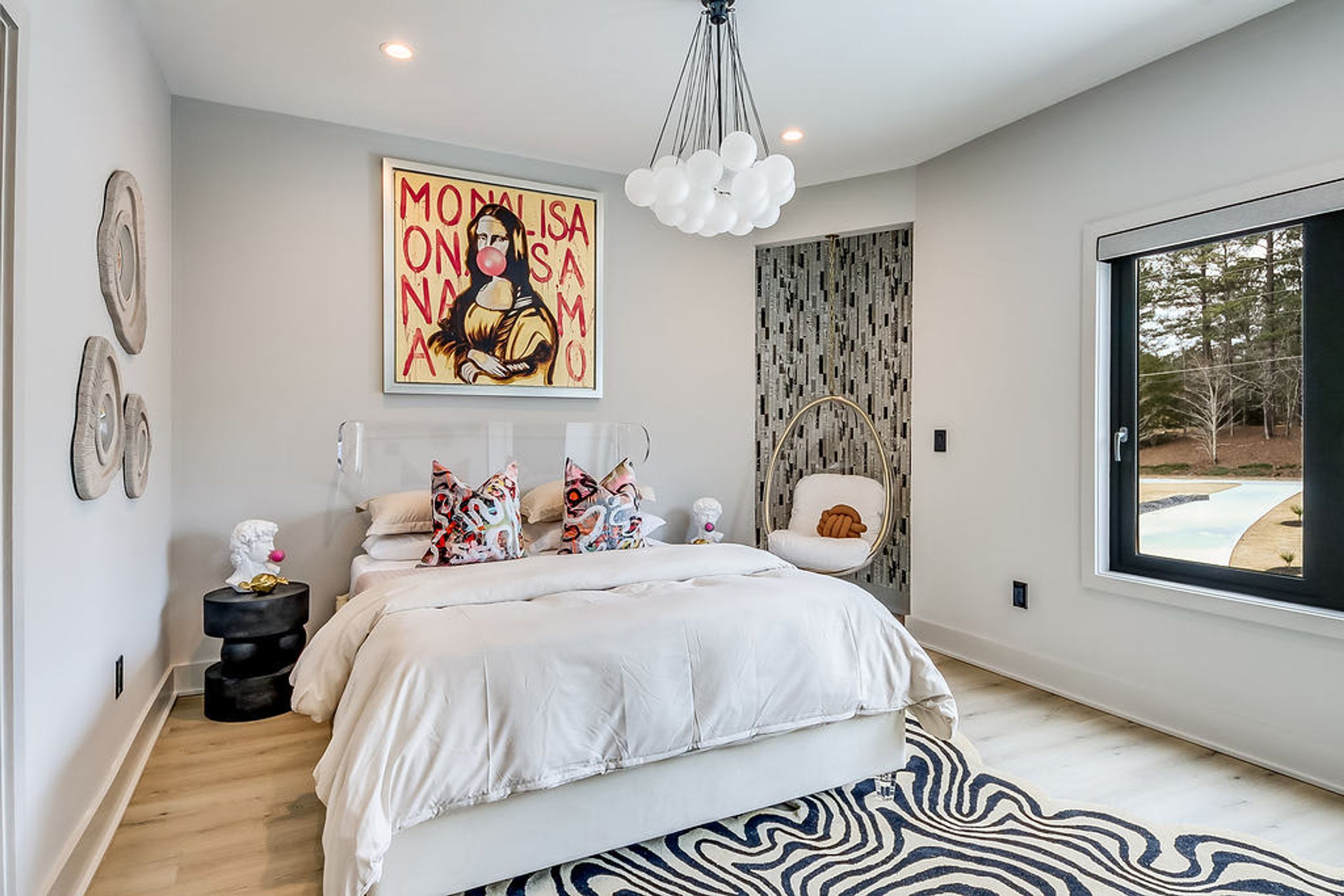 A modern bedroom with a large bed, a unique wall art piece, a hanging light fixture, and a view of the outdoors through a large window.