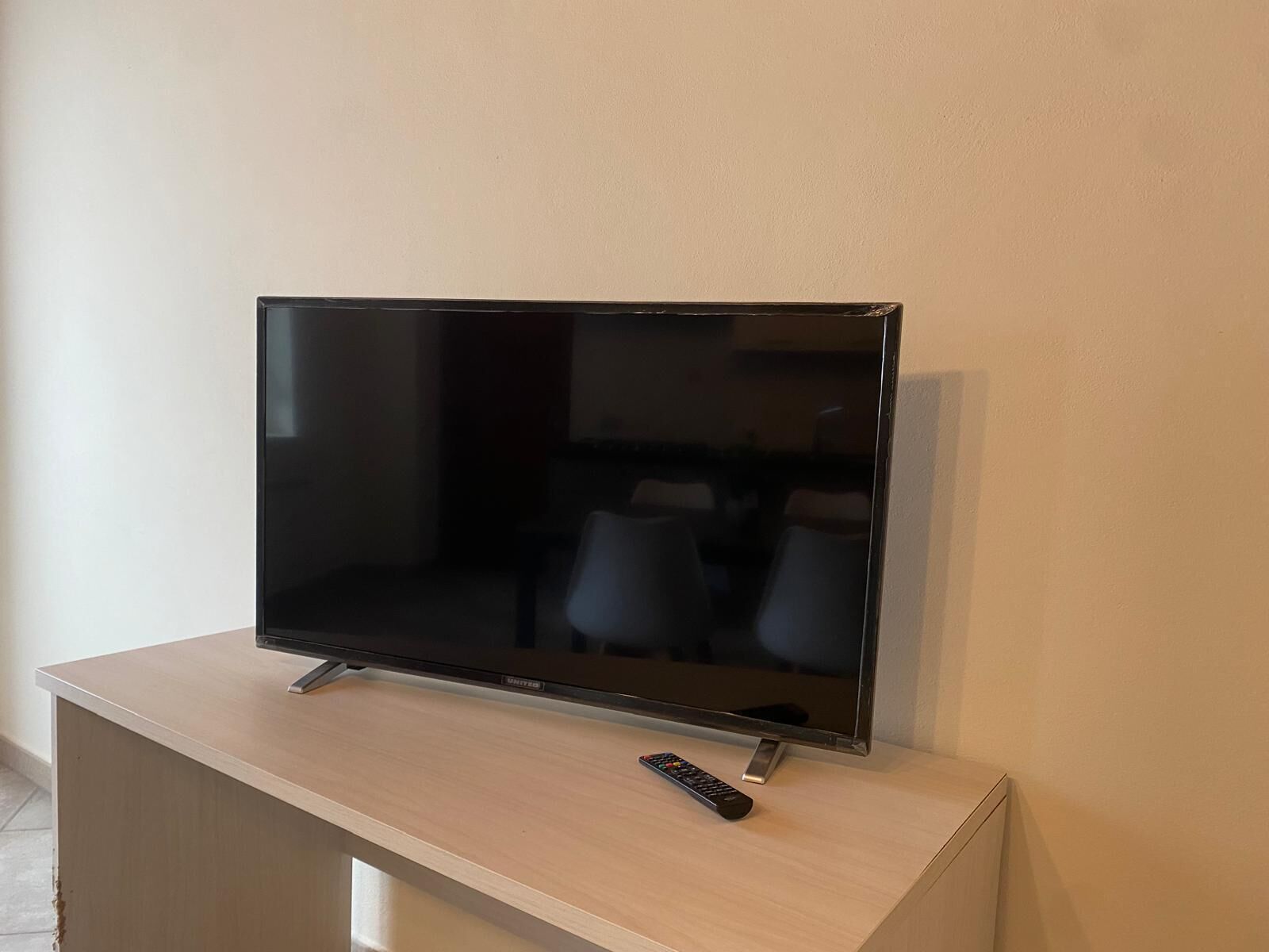A television on a wooden stand in a room with a plain wall and a glimpse of a dining area in the background.