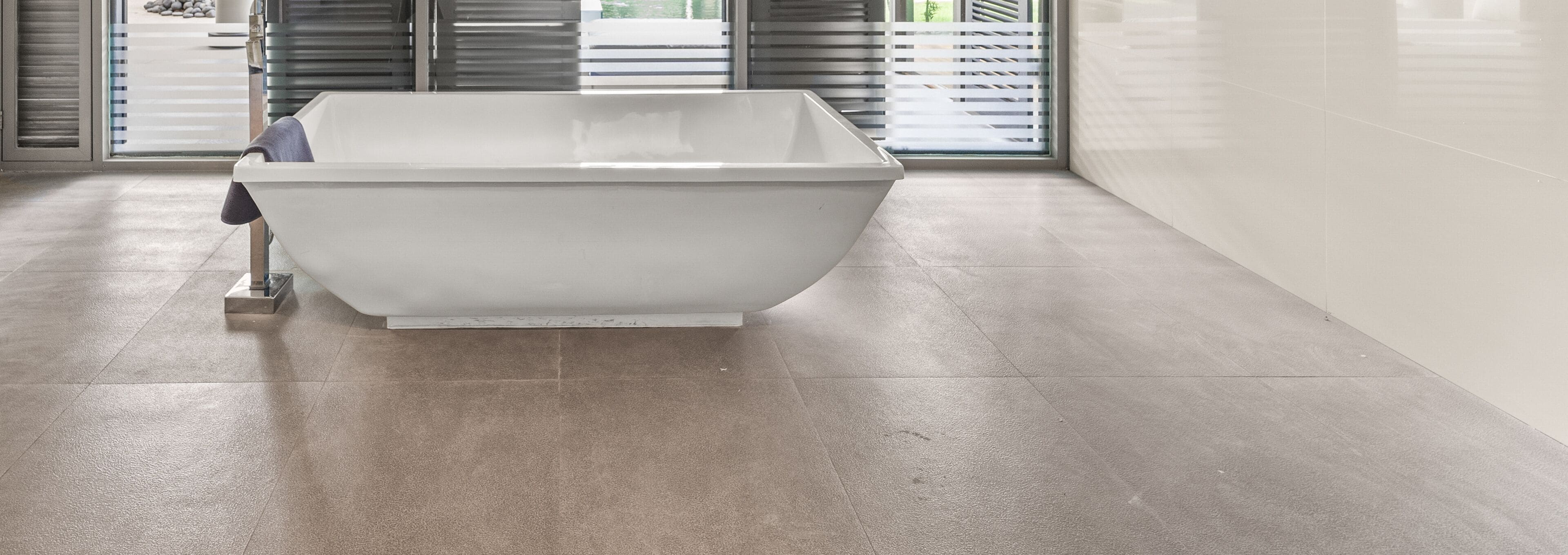 A modern bathroom with a freestanding bathtub, large windows with blinds, and a tiled floor.