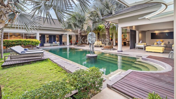 Un área de piscina interior moderna con techo de vidrio, palmeras y un espacio de relax con un sofá amarillo.