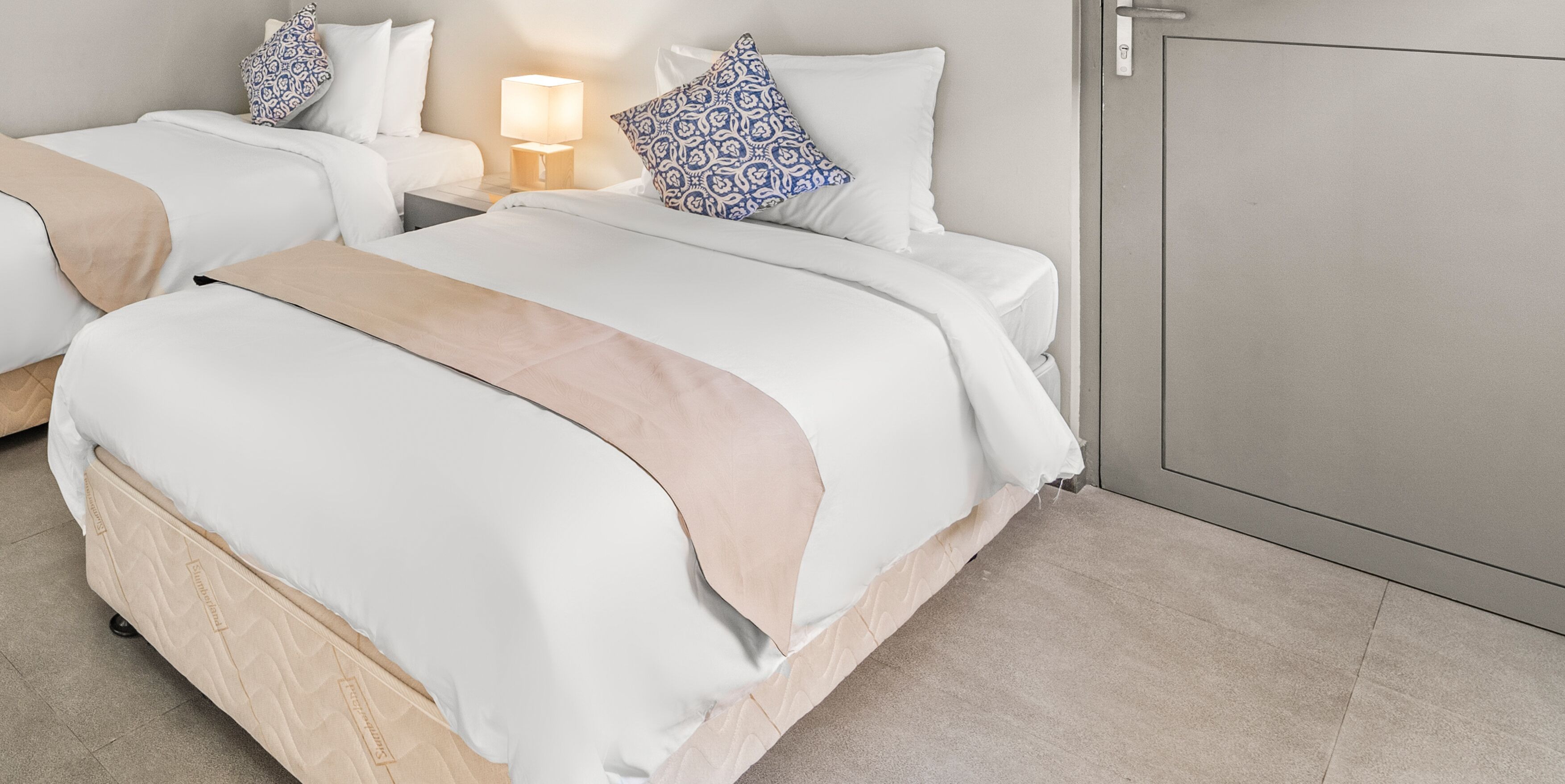 A hotel room with two beds, a nightstand with a lamp, and a wardrobe.