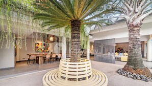 Un espacio interior moderno con una palmera central rodeada por un banco circular, un comedor con sillas y una zona de estar con cojines.